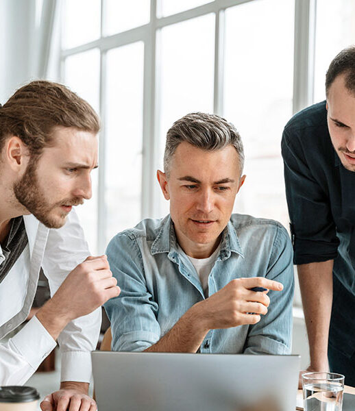 Three men collaborating intently over a laptop in a bright office. Three men collaborating intently over a laptop in a bright office.