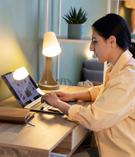 Woman working on a laptop at a wooden desk with a lamp and plant. Woman working on a laptop at a wooden desk with a lamp and plant.