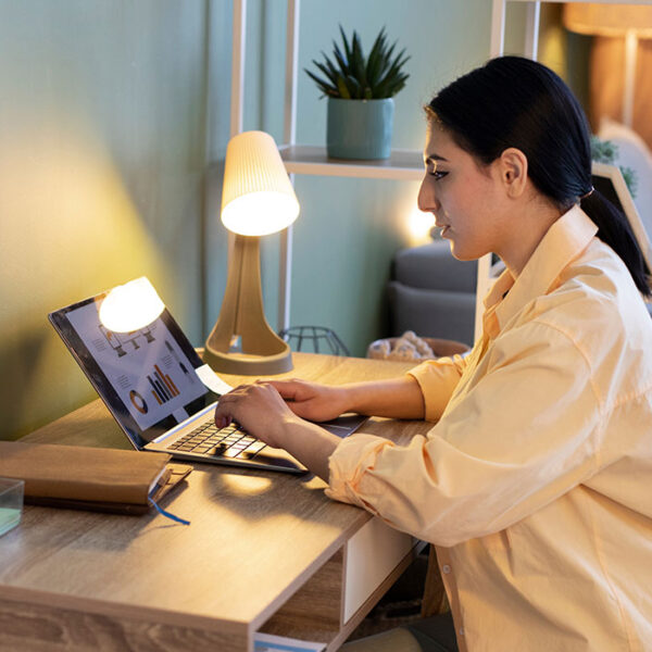 Woman working on a laptop at a wooden desk with a lamp and plant. Woman working on a laptop at a wooden desk with a lamp and plant.